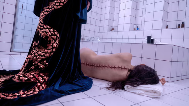 a still from "the substance" in which a figure in a robe with a snake on it stands over a naked woman with rough stitches up her spine, on the floor of a stark white-tiled bathroom