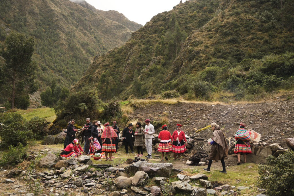 People standing in a clearing in the Peruvian mountains, some are wearing red traditional garb. They are beside a dry rocky stream bed
