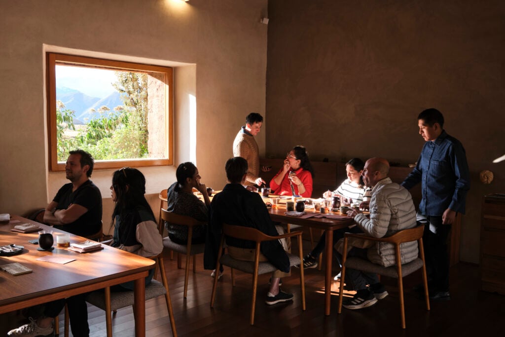 People sharing a meal, sitting around a wooden table, with wooden chairs. Outside a window is a view of the Peruvian mountains