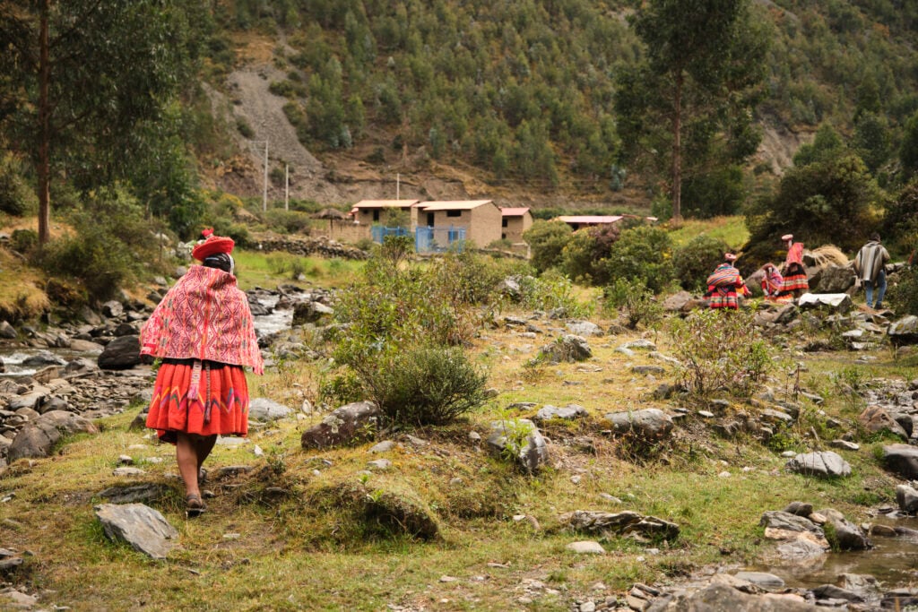 Women in brightly colored garb, along side a rock stream shore, mountains in background
